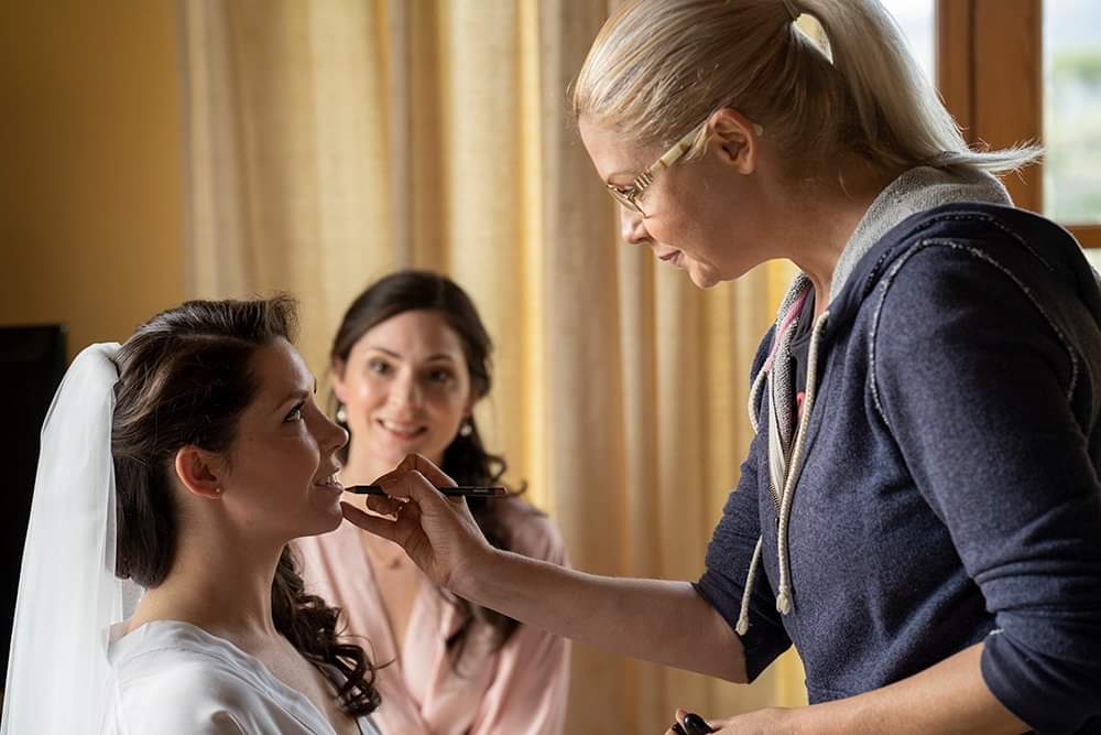 Makeup artist Arienne Aereboe applies makeup to a bride wearing a veil, while another woman watches attentively in the background.