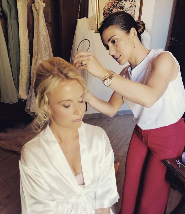 Stylist Suela works on a bride’s hair, styling it into an elegant updo as the bride sits in a white robe.